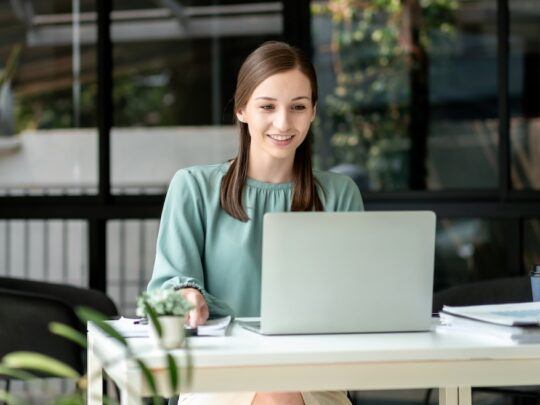 An adult smiling woman works on her computer from her home office. She is a European entrepreneur working with technology for business and e-commerce.